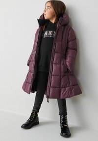 Lång, puffig rock i djupt lila med hög krage, bärs över en svart hoodie och svarta byxor, tillsammans med blanka svarta snörboots.