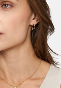 Goldene Creolen mit einem wellenförmigen Design, verziert mit kleinen klaren Steinen. Glatte Textur und eine schlanke Kettenhalskette sichtbar.