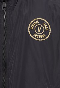 Črna bomberska jakna z okroglo zlatim vezenim logotipom "Versace Jeans Couture." Zapiranje z zadrgo s teksturirano tkanino.