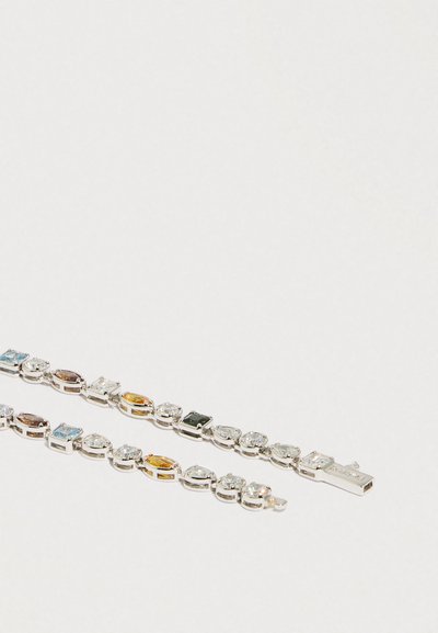 Dwie srebrne bransoletki z naprzemiennie ułożonymi przezroczystymi, niebieskimi, żółtymi i ciemnozielonymi prostokątnymi i kroplowymi kamieniami szlachetnymi na białym tle.