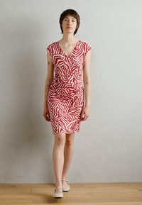 Robe à motifs rouges et blancs avec une silhouette ajustée, un décolleté en V et des manches courtes. Texture lisse, longueur genoux, portée avec des chaussures de couleur claire.