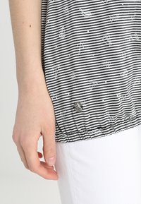 Vänster hand vilar bredvid kroppen klädd i en svart- och vitrandig skjorta med små segelbåtsmönster och vita byxor.