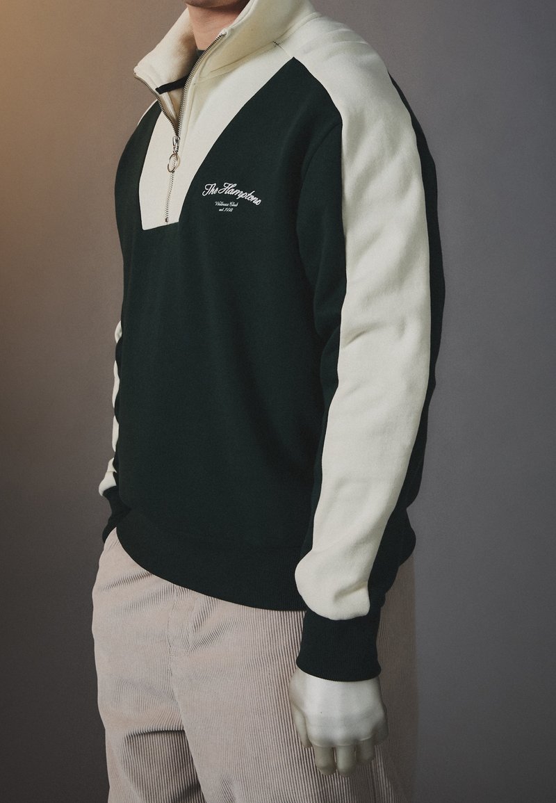 Zwart en crèmekleurige quarter-zip pullover sweatshirt met een V-vormig ontwerp, voorzien van geborduurde tekst en contrasterende mouwpanelen.