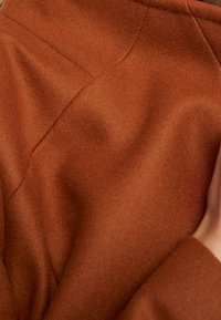 Tejido de mezcla de lana de color óxido con una superficie suave y texturizada, que presenta detalles de costura prominentes y un corte moderno.