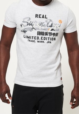 Mężczyzna w białej limitowanej koszulce SuperDry z grafiką oraz czarnych spodniach, stojący z rozluźnionymi ramionami wzdłuż ciała.