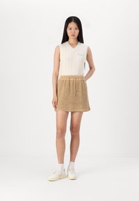 Lys kremfarget ermeløs strikket topp med v-hals og blå broderi, sammen med en teksturert beige strikket miniskjørt og hvite sneakers.