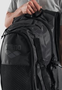Mochila negra de nylon con panel de malla texturizada, múltiples cremalleras y detalles de logo. Cuenta con un mango acolchado y correas ajustables.