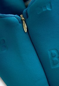 Tirkizna jakna sa patentnim zatvaračem napravljena od teksturiranog materijala, s zlatnim patentnim zatvaračem i reljefnim detaljem "B". Glatka završna obrada s prilagođenim dizajnom.