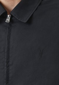 Zwarte jack met ritssluiting, puntige kraag en tonale geborduurde dieren schedel logo op de borst. Glad stofmateriaal.