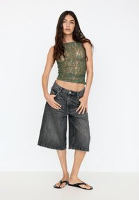 Green lace crop top with ruched detailing, paired with loose black denim culottes. Model wears black flip-flops, standing against a white backdrop.