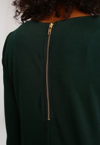 Mörkgrön plagg med en slät yta, med en guld dragkedjedetalj längs ryggen, ovanför halsringningen.