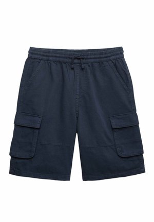 Junge trägt ein hellrosa übergroßes T-Shirt, marineblaue Cargo-Shorts und beige Slip-On-Schuhe, steht mit den Händen in den Taschen und geneigtem Kopf.