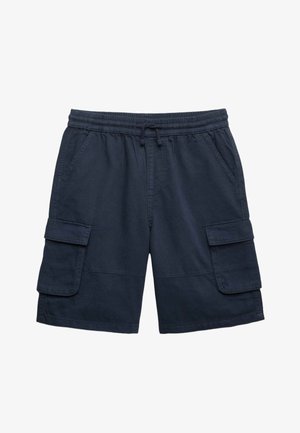 Chłopiec ubrany w jasnoróżowy, oversize'owy T-shirt, granatowe szorty bojówki oraz jasnobrązowe wsuwane buty, stoi z rękami w kieszeniach i przechyloną głową.