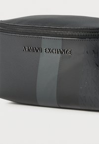 Μαύρο υφασμάτινο πουγκί με φερμουάρ, με μεταλλικό λογότυπο «ARMANI EXCHANGE» και κάθετη ματ ρίγα στο μπροστινό μέρος.