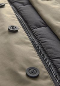 Gros plan sur un tissu de veste beige avec des boutons noirs et une fermeture éclair noire, à côté d'une doublure noire matelassée.