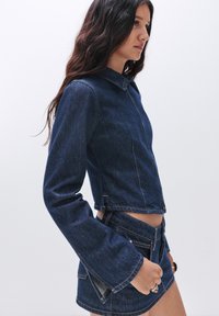 Giacca di denim blu scuro con un design accorciato, colletto appuntito, maniche lunghe e svasate, e dettagli di cucitura visibili. Abbinata a una gonna coordinata.