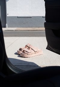 Sandales compensées rose clair avec des lanières sur un trottoir en béton ensoleillé à l'extérieur d'une porte de voiture ouverte près d'une rue.