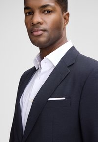 Dunkelblaues Anzug mit strukturiertem Stoff, kombiniert mit einem weißen Hemd und einer Tasche. Der Kragen ist strukturiert und die Passform ist tailliert.