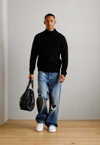 Pull en maille noir avec fermeture éclair, jeans déchirés bleu clair, baskets blanches et sac en cuir noir. Le modèle se tient devant un fond neutre.
