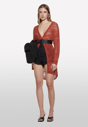 Un cardigan tricoté rouge rouillé avec un décolleté en V profond, porté sur un short noir taille haute. Un chapeau noir tissé et des sandales plates noires complètent l'ensemble.
