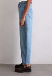 Lyseblå høytlivsjeans med rett ben, med en lett falming. Kombinert med mørkebrune loafers, satt mot en nøytral bakgrunn.