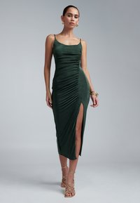 Smaragdgrünes, gerafftes Kleid mit Spaghettiträgern, hohem Schlitz und weichem, strukturiertem Stoff. Kombiniert mit goldenen Armbändern und Riemchensandalen.