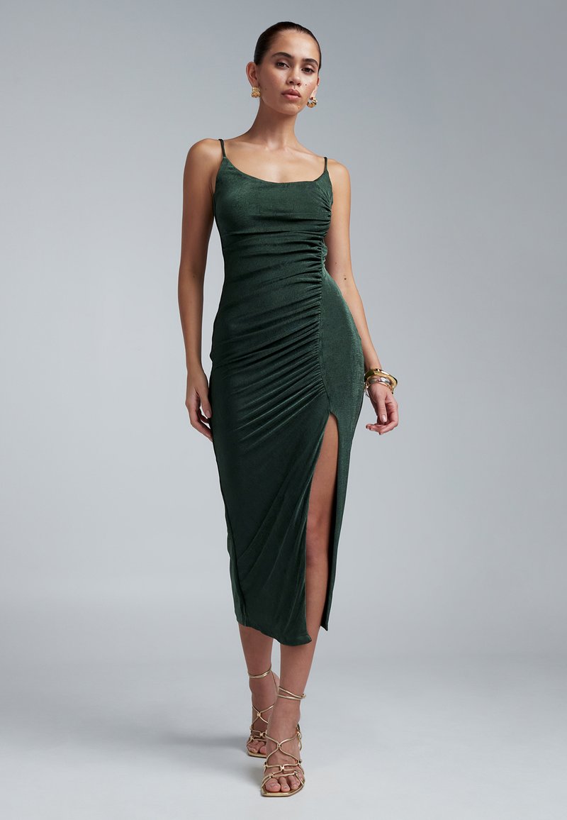 Smaragdgrünes, gerafftes Kleid mit Spaghettiträgern, hohem Schlitz und weichem, strukturiertem Stoff. Kombiniert mit goldenen Armbändern und Riemchensandalen.