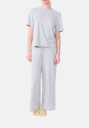 Vrouw draagt een lichtgrijze top met korte mouwen en wijde broek met subtiele horizontale strepen, staand met voeten dicht bij elkaar op een witte achtergrond.