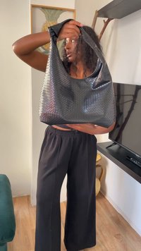 Person holding a large black textured handbag, standing indoors near a TV and pineapple artwork, wearing black pants and a black top.
