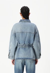 Lys blå denimjakke med avslappet passform, knappet krage og belte i midjen. Har overdrevne ermer og vertikale sømdetaljer.