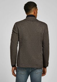 Man wearing a brown textured blazer and blue jeans, standing with his back to the camera against a plain background.