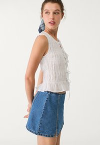 Weißes, strukturiertes Oberteil mit Schnürung vorne und Peplum-Saum, kombiniert mit einem hoch taillierten, blauen Denim-Minirock mit Rückenschlitz.