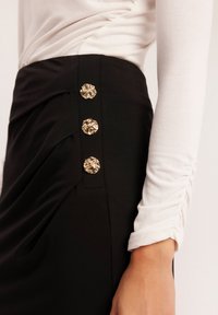 Czarna spódnica o drapowanej fakturze z trzema dekoracyjnymi złotymi guzikami po boku, zestawiona z dopasowaną białą bluzką z długim rękawem.