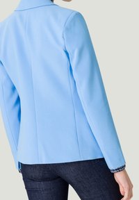 Blazer bleu clair au design ajusté, fabriqué en tissu lisse et doté d'un col. Les accents incluent un détail de poignet à motifs.