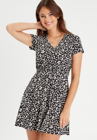 Schwarzes Kleid mit weißem Blumenmuster, kurzen Ärmeln, V-Ausschnitt und Bindeband in der Taille. Besteht aus leichtem, weichem Stoff und hat eine ausgestellte Silhouette.