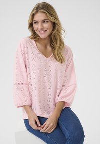 Blusa rosa con scollo a V e maniche a tre quarti, caratterizzata da motivi floreali ritagliati e polsini elasticizzati, abbinata a jeans blu.