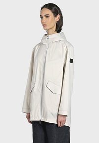 Giacca impermeabile beige chiaro con cappuccio, chiusura con zip, due tasche anteriori e un piccolo logo nero sulla manica. Tessuto liscio, vestibilità rilassata.