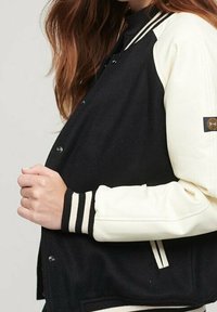 Svart och krämfärgad varsityjacka med ullkropp och läderslevar. Har randiga ribbade manschetter och en logopatch på ärmen.