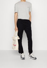 Grå kortærmet t-shirt, sorte tætsiddende bukser og multicolore sneakers. Holder en beige tote bag med rød og grå tekst.