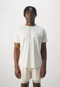 Camiseta deportiva en un tono beige claro con mangas cortas, cuello redondo y un pequeño logo negro; textura suave y silueta ajustada.