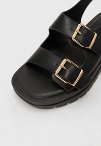Musta sandaali, jossa on kaksi leveää remmiä kultaisilla soljilla, teksturoitu pohja ja paksu musta pohja. Elegantti ja toimiva muotoilu.