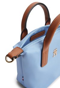 Ανοιχτό μπλε τσάντα χειρός με λαβές από δέρμα σε καφέ απόχρωση, μεταλλικό λογότυπο στο μπροστινό μέρος, φερμουάρ στην κορυφή και μικρή λεπτομέρεια με σημαία στη ραφή του πλαισίου.