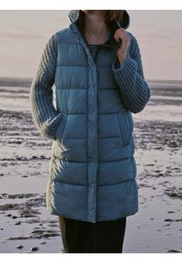 Padded teal coat with grey knit sleeves, featuring a high collar, front pockets, and a zipper closure. Soft texture with a quilted pattern.