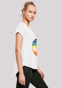 White short-sleeve T-shirt with a distressed multicolored alien graphic in orange, blue, and teal. Curved hemline, relaxed fit.