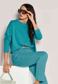 Türkiser gestrickter Pullover mit weitem Ausschnitt, kombiniert mit passenden figurbetonten Hosen. Das Model trägt übergroße braune Sonnenbrillen und hält eine leichte Tasche.