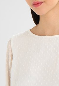 Weiße Bluse mit rundem Ausschnitt, transparenten langen Ärmeln, strukturiertem Stoff und erhabenem Pünktchenmuster in der gesamten Bluse.