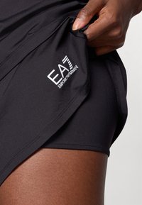 Černé sportovní šortky s těsným vnitřním vrstvou. Obsahují vyšívané bílé logo EA7 na vnější straně a mají hladký povrch.