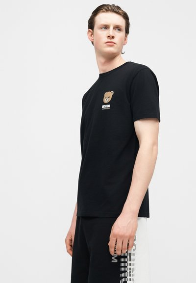 Νέος άνδρας που φοράει μαύρο μπλουζάκι Moschino με λογότυπο αρκουδάκι και μαύρο-άσπρο σορτς με ριγέ κείμενο στο πλάι.