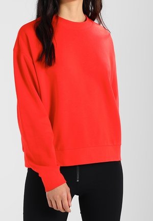 Sudadera roja de manga larga, con cuello redondo, un corte holgado y textura suave, combinada con pantalones negros ajustados.