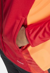 Rote und orange Jacke mit glatter Textur, ausgestattet mit einer Reißverschlusstasche an der Seite und 'AEROREADY'-Branding am unteren Rand.